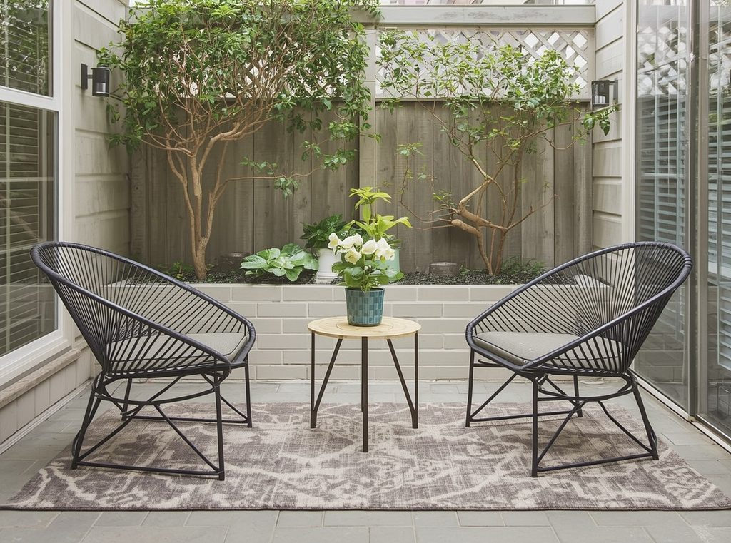 A compact patio seating area anchored by a neutral outdoor rug beneath two chairs and a small round table